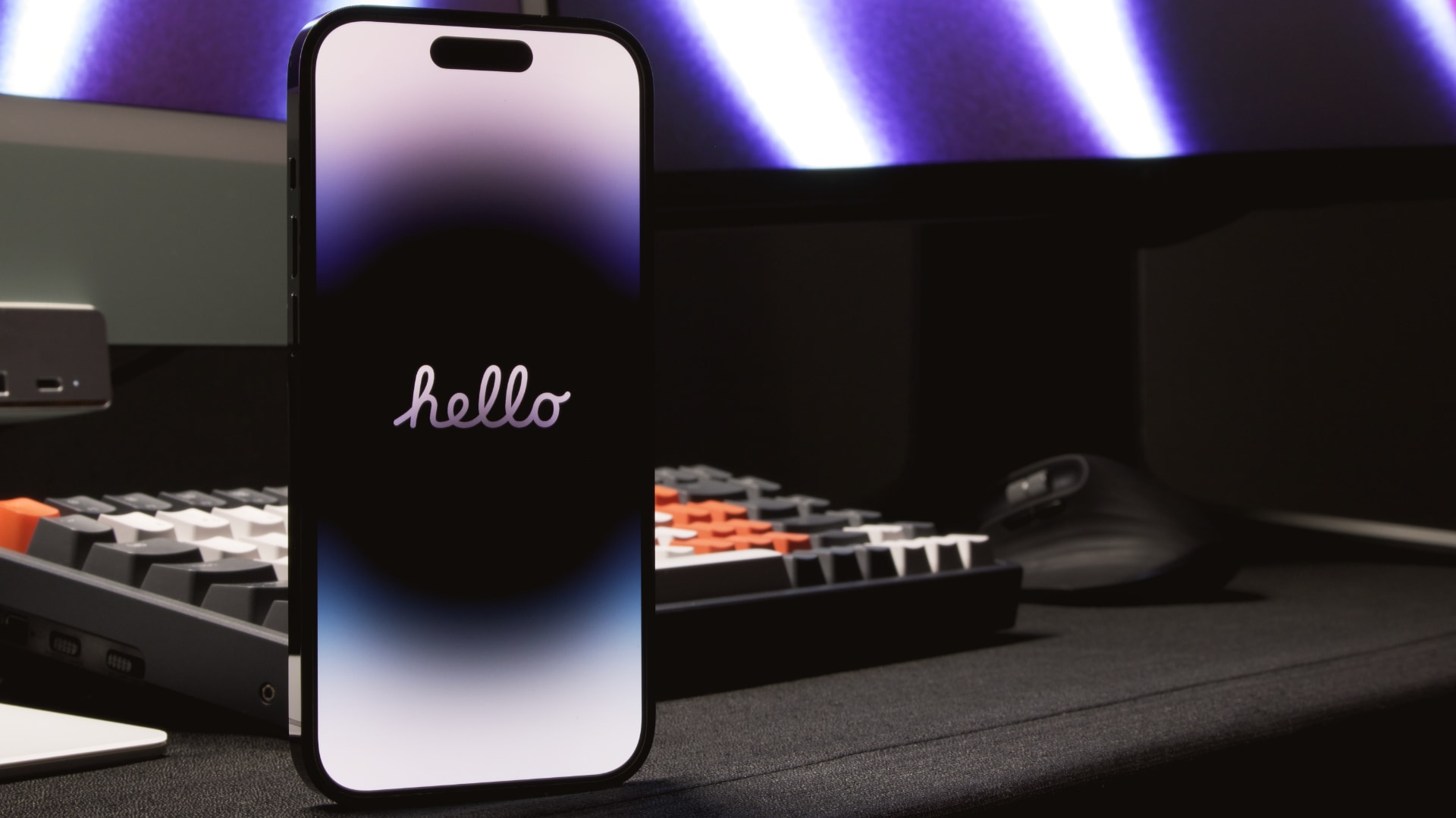 iPhone 14 Pro Max zeigt einen Hallo -Bildschirm an, der vor einem Computermonitor und einer mechanischen Tastatur auf einem Holzschreibtisch aufrecht steht.